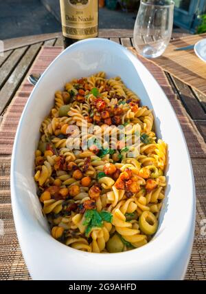 Nahaufnahme von Nudelgerichten: Fusilli mit Kichererbsen, Kapern, Oliven und Tomaten in weißer Auflaufform auf dem Freilufttisch bei Sonnenschein mit Weinflasche serviert Stockfoto