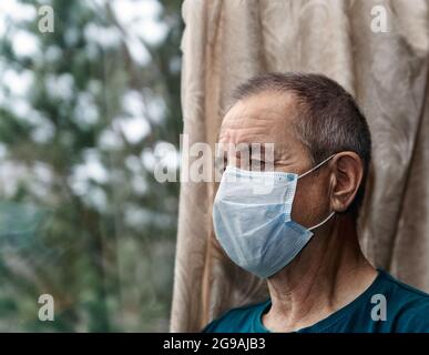 Älterer Mann mit Maske aus Covid 19 steht am Fenster. Selbstisolationsmodus. Stockfoto