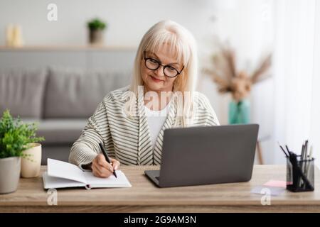 Reife Dame, die im Notebook schreibt und zu Hause einen Laptop benutzt Stockfoto