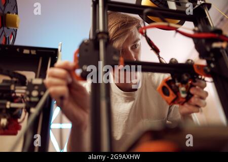 Niedriger Winkel des jungen Mannes Befestigungsmechanismus des 3D-Druckers während der Arbeit in der Werkstatt Stockfoto