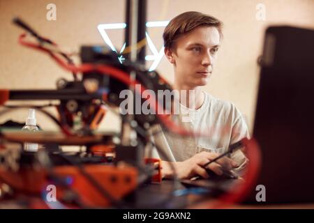 Junger männlicher Ingenieur, der während der Arbeit im Studio am Tisch sitzt und Netbook in der Nähe des 3D-Druckers durchstöbert Stockfoto