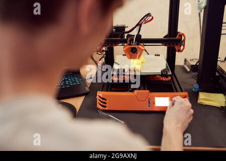 Hoher Winkel anonymer männlicher Einstellregler auf 3D-Drucker bei der Erstellung eines Plastikmodells in der Werkstatt Stockfoto