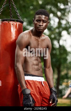 Muskulöser schwarzer Profi-Boxer schwitzt in Handschuhen beim Boxtraining im Freien, junger afroamerikanischer Boxer beim Training auf dem Straßenspielplatz. Stockfoto