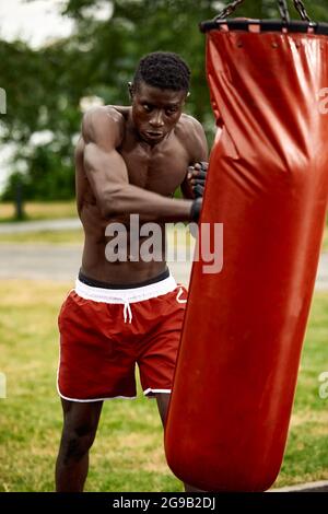 Muskulöser schwarzer Profi-Boxer schwitzt in Handschuhen beim Boxtraining im Freien, junger afroamerikanischer Boxer beim Training auf dem Straßenspielplatz. Stockfoto