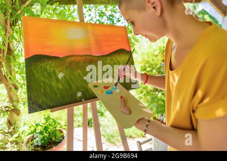 Junge Künstlerin arbeitet an ihrer Kunst Leinwand Malerei im Freien in ihrem Garten. Krebs-Überlebende, Achtsamkeit, Kunsttherapie, Kreativitätskonzept. Stockfoto