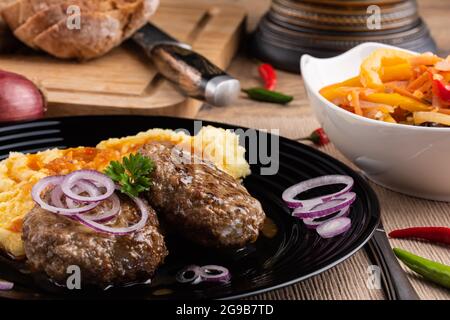 Koteletts mit zerkleinerten Kartoffeln, mit Sauce bestreut und mit gehackten Zwiebeln bestreut. All dies auf einem schwarzen Teller, Salat in einer weißen Tasse, auf dem Abendessen Stockfoto