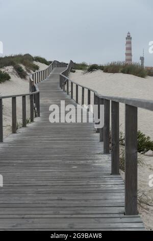 Hölzerne Gehwege entlang der Dünen des Strandes von Barra mit dem Leuchtturm von Aveiro im Hintergrund an einem bewölkten Tag. Portugal Stockfoto