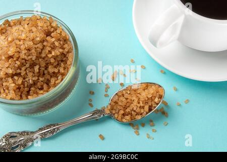 Bernsteinfarbene Zuckerkristalle in einem Löffel in der Nähe einer Glaszuckerschale und eine Tasse heißes Getränk auf blauem Hintergrund. Unraffinierter brauner Rohrzucker für Kaffee und Tee Clo Stockfoto