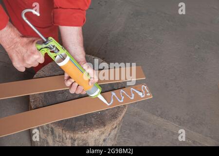 Die Hände der Arbeiter in einem roten Overall Holding eine Spritze, die Waffe für das Auftragen von Leim. Ein Arbeitnehmer gilt Weißleim in Streifen von laminierten Holzfaserplatten, lami Stockfoto