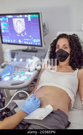 Junge Schwangere mit Gesichtsschutzmaske aufgrund der Coronavirus-Pandemie, covid 19 bekommen einen Ultraschall, um ihr Baby in einer Klinik zu sehen Stockfoto