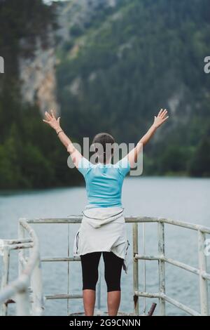 Die reife Frau steht und hebt die Arme und blickt auf einen blauen See mit großen Bergen davor. Konzept von Reisen und Freiheit Stockfoto