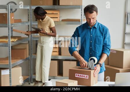 Porträt eines jungen Mannes, der im Lager mithilfe eines Kassettenspenders Kartons mit zerbrechlichen Aufklebern verpackt und Platz für Kopien verwendet Stockfoto