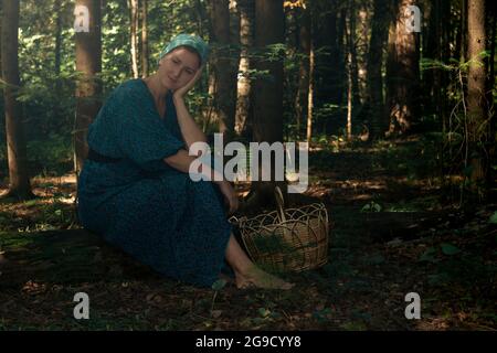 Die junge Frau in volkstümlicher Bauernkleidung mit einem Korb zum Pflücken wilder Pflanzen sitzt auf einem Baumstamm im Nadelwald Stockfoto