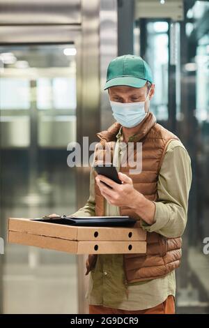 Vertikales Porträt des Lieferers, der eine Maske trägt und Pizza hält, während er im Aufzug steht Stockfoto