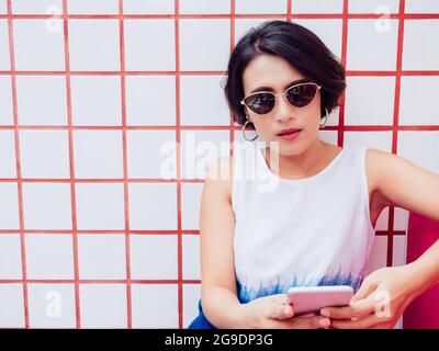 Attraktive stilvolle asiatische Frau kurze Haare tragen weißes Hemd und Sonnenbrille sitzen auf rotem Sitz und mit Smartphone auf weißen und roten Fliesen Wandrückseite Stockfoto