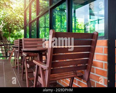 Leere rote gemütliche Holzstühle und ein runder Couchtisch aus Holz in der Nähe des Glasfensters und der Ziegelwand mit grünem Garten im Café, im Sommer. Stockfoto