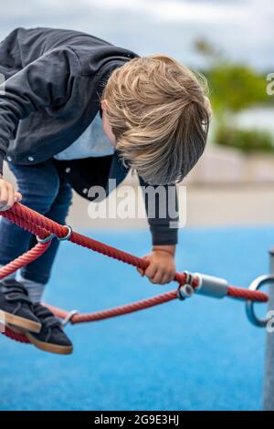 Der kleine Junge spielt begeistert mit einem gedehnten Seilnetz auf dem Spielplatz, um das Gleichgewicht des Kindes zu entwickeln und die motorischen Fähigkeiten zu entwickeln, die ihm die Möglichkeit geben Stockfoto