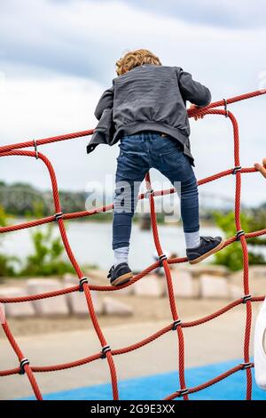 Der kleine Junge spielt begeistert mit einem gedehnten Seilnetz auf dem Spielplatz, um das Gleichgewicht des Kindes zu entwickeln und die motorischen Fähigkeiten zu entwickeln, die ihm die Möglichkeit geben Stockfoto