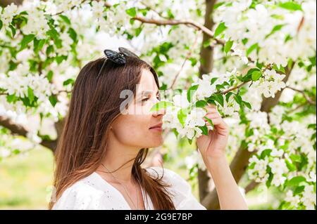 Lächelnd und glücklich posiert das Weibchen in weißen Kleidern. Fröhliche hübsche Frau auf Apfelbaum Hintergrund. Junge kaukasische Frau. Attraktiv und b Stockfoto