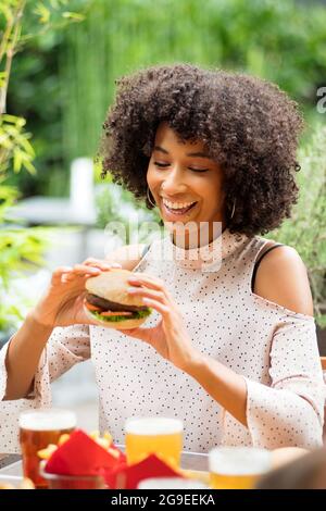 Lebhaft glückliche junge schwarze Frau, die einen Hamburger in ihren Händen mit einem strahlenden Lächeln und einem Blick der Vorfreude in einem Restaurant im Freien isst Stockfoto