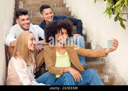 Eine Gruppe von glücklichen jungen Freunden, die auf einer Außentreppe für ein Selfie auf einem Mobiltelefon posieren, lachen und lächeln an der Kamera Stockfoto