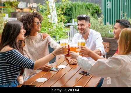 Eine Gruppe verschiedener junger Freunde, die in einem Open-Air-Restaurant oder Pub feiern und sich gegenseitig mit kalten Bieren toasten, während sie lachen und lächeln Stockfoto