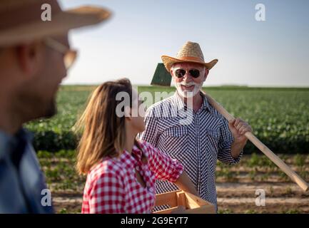 Junger Bauer im Vordergrund mit Hut und Sonnenbrille. Leitender Landwirt, der mit einer Gartenhacke lächelnd auf der Schulter steht. Junge Frau, die dazwischen steht Stockfoto