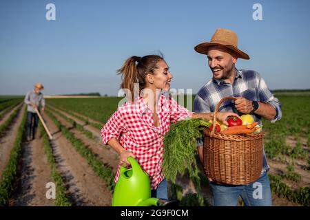 Junger Mann hält Korb voller Gemüse lächelnd. Junge Frau mit Gießkannen, die Tomate berührt und spricht. Alter Mann mit Gartenhacke im Feld i Stockfoto