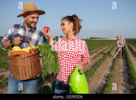 Senior Farmer jäten mit Gartenhacke im Hintergrund. Junge Frau hält Gießkanne und Tomate Blick auf jungen Mann mit Korb voller Vegetabl Stockfoto