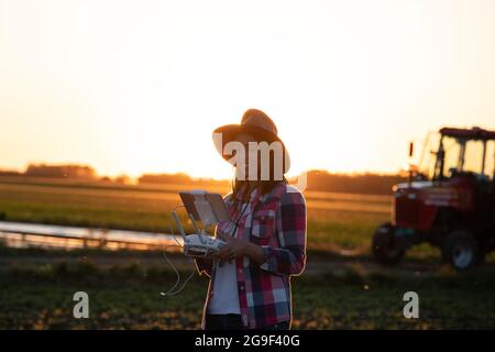 Junge Agrarwissenschaftlerin vermessung Land mit Drohne bei Sonnenuntergang. Landwirt, der mit innova auf dem Feld vor dem Traktor steht und einen Drohnencontroller mit Tablet hält Stockfoto