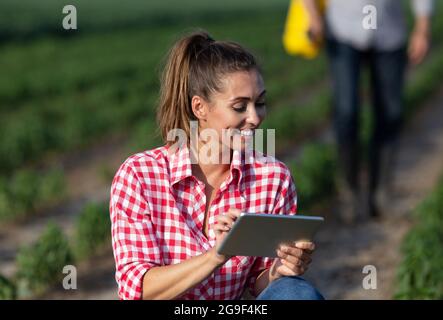 Attraktive junge Farmer hocken auf dem Feld mit Tablet lächelnd. Bauer läuft im Hintergrund und arbeitet mit einem tragbaren Handsprüher. Stockfoto