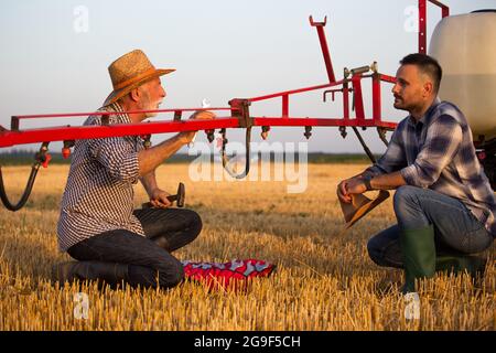 Junger Bauer hockend mit Strohhut in der Hand und hört zu. Älterer Landwirt, der Schlüssel und Hammer hält, um die Auslegerspritze am Traktor zu reparieren, Explainin Stockfoto