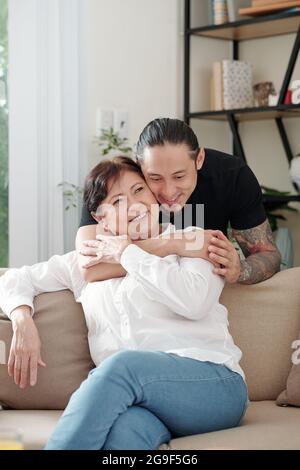 Älterer Sohn umarmt seine glückliche Mutter von hinten, während sie auf dem Sofa im Zimmer sitzt Stockfoto