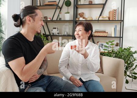 Glückliche Mutter trinkt Tee und lacht während eines Gesprächs auf dem Sofa mit ihrem Sohn im Wohnzimmer Stockfoto