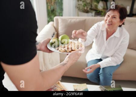 Nahaufnahme eines Mannes, der ein Tablett mit Keksen und Obst hält und eine Frau behandelt, die auf dem Sofa im Zimmer sitzt Stockfoto