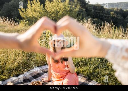 Blick durch das Herzzeichen, hergestellt mit Händen von zwei Frauen. Ziemlich sympathisch lächelnde junge Frau in Kleid und Hut, die draußen auf einer karierten Decke auf einer wunderschönen Wiese sitzt und die Kamera anschaut Stockfoto