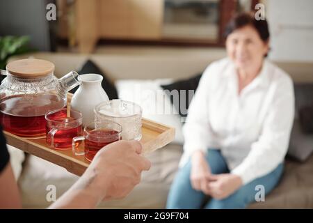 Nahaufnahme eines Sohnes, der ein Tablett mit Teekannen und Tassen hält und es seiner Mutter serviert, während sie auf dem Sofa im Zimmer sitzt Stockfoto
