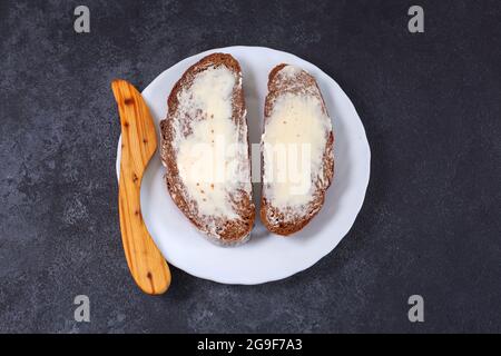 Zwei Stück handwerkliches Brot mit Butter und Holzmesser auf einem weißen Teller. Draufsicht. Stockfoto