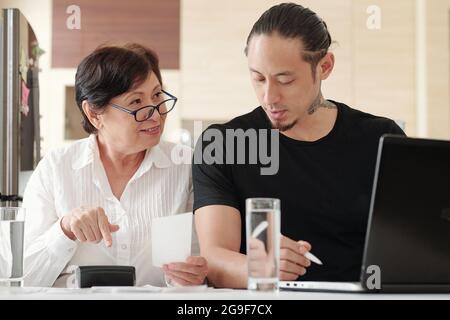Junger Mann, der am Tisch sitzt und mit einer reifen Frau am Laptop arbeitet, die in der Nähe sitzt, indem er mit der Finanzabteilung arbeitet und mit dem Mann darüber diskutiert Stockfoto