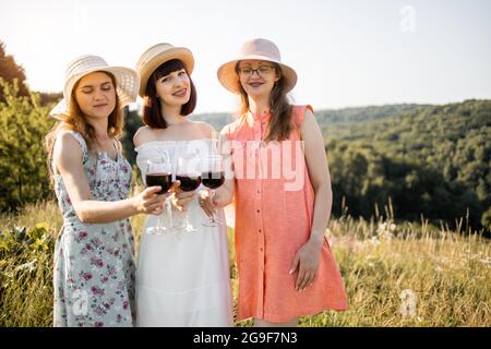 Drei schöne Freundinnen klirren mit einem Glas Rotwein, während sie auf dem Gras stehen und an sonnigen Tagen auf der Sommerwiese gemütlich picknicken, ganz in der Nähe Stockfoto