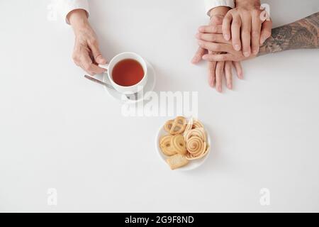 Aus der Perspektive der Mutter, die am Tisch sitzt und die Hand ihres Sohnes hält, während sie Tee mit Keksen trinkt Stockfoto