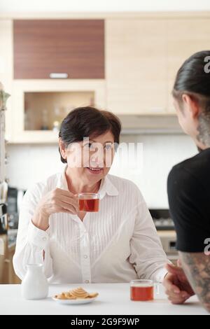 Asiatische reife Frau sitzt am Tisch, trinkt Tee und spricht mit ihrem Sohn in der Küche Stockfoto