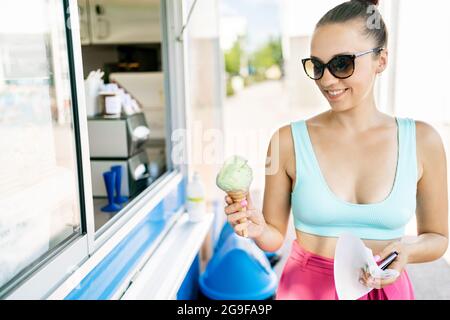 Zufriedener Kunde, der Eis aus dem Salon, LKW, Lieferwagen oder Kiosk kauft. Im Sommer Eiscreme zum Mitnehmen. Lächelnde Frau, die Eiscreme in der Hand hält. Stockfoto