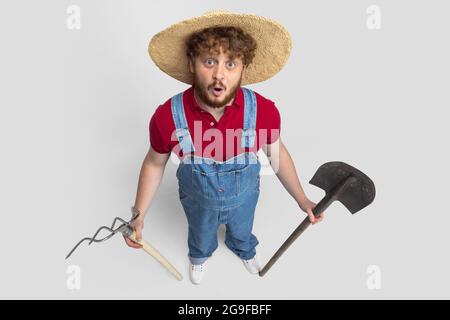 Hochwinkelansicht des rothaarigen bärtigen Mannes, Landwirt in Strohhut mit Gartengeräten isoliert über weißem Studiohintergrund. Konzept der professionellen Stockfoto