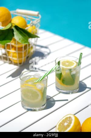 Hausgemachte Limonade in Gläsern mit Zitronenscheibe, Zitronenkorb, Trinkhalm, auf weißem Tisch, am Rand des Pools mit türkisblauem Wasserhinterm Stockfoto