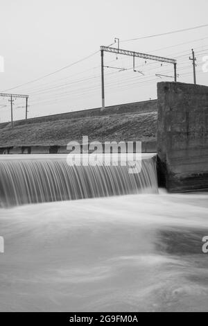 Der Unterschied in der Höhe im Betonkanal neben den Gleisen der Eisenbahn. Schwarzweißfotografie mit Langzeitbelichtung. Stockfoto