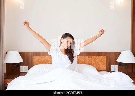Porträt einer schlaflich lächelnden jungen Frau, die nach dem Aufwachen in ihrem Bett sitzt und sich ausdehnt Stockfoto