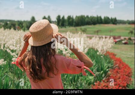 Frau in Hut in der Nähe von Blumenbeet. Mädchen in rosa Kleid, in Strohhut steht mit ihrem Rücken zur Sonne in floralen weißen und roten Feld, hält den Hut Stockfoto