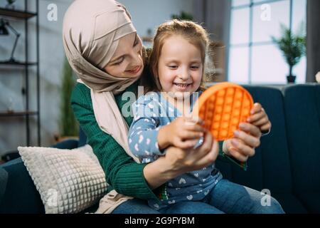 Pädagogische Spiele für Kinder, poppit, antistressige sensorische Pop-it-Spielzeug. Aufgeregt fröhlich entzückendes kleines Mädchen, spielt ein beliebtes Anti-Stress-Spiel mit Knöpfen, sitzt auf dem Sofa mit ihrer arabischen Mutter in Hijab Stockfoto