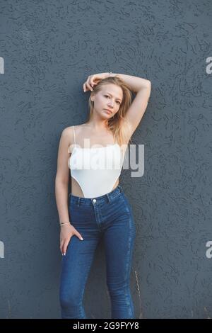 Junge blonde Frau im weißen ärmellosen T-Shirt und blauen Jeans, die über grauem Wandhintergrund stehen Stockfoto